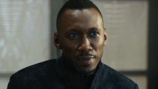 Mahershala Ali sits with a sly smile and glowing blue eyes in Alita: Battle Angel.