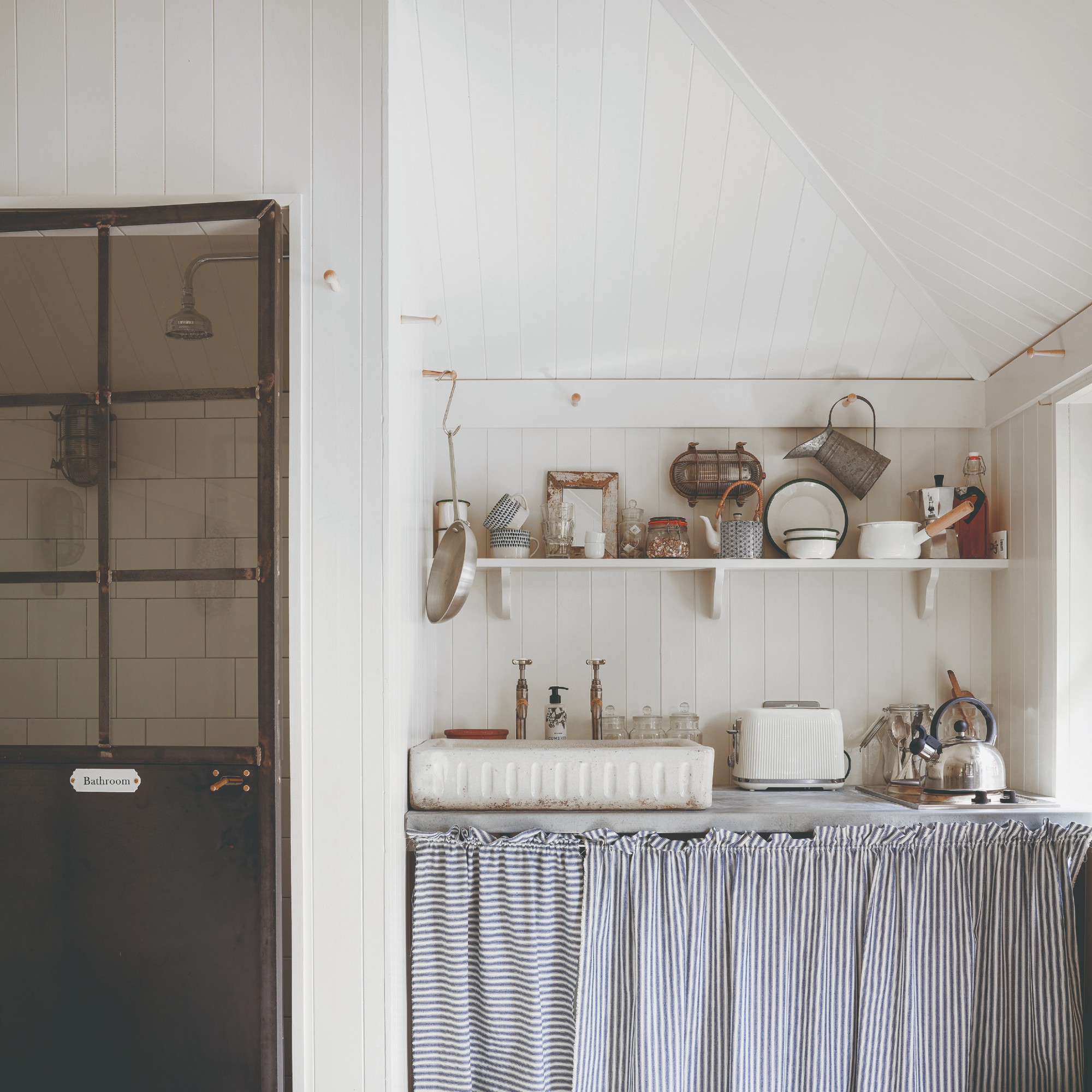 A rustic-style wall-panelled kitchen painted in white with a striped blue curtain covering the under-sink area