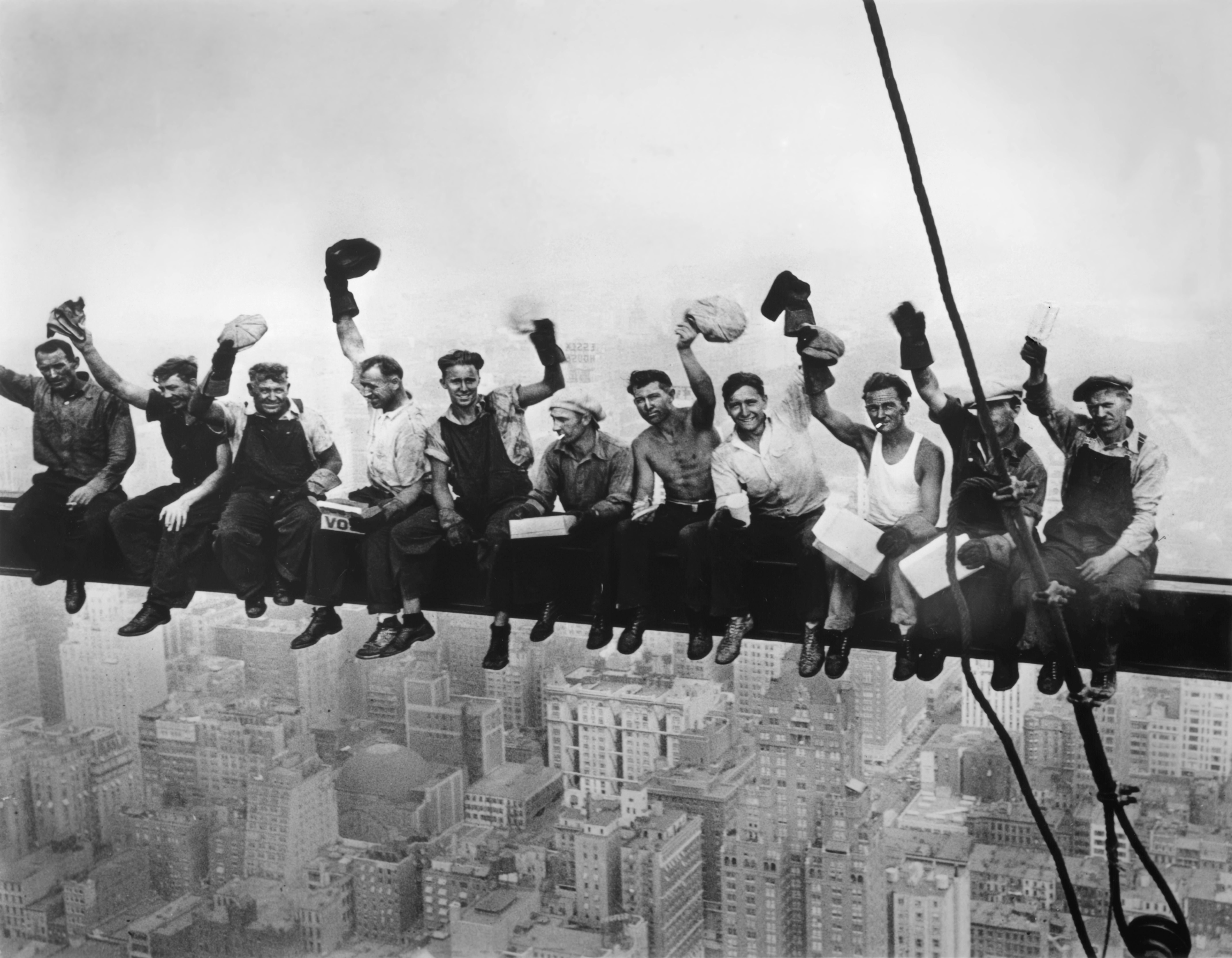The same eleven ironworkers on their beam, this time caught mid-cheer, hats raised aloft and faces grinning broadly at the camera against the same vertiginous Manhattan backdrop.