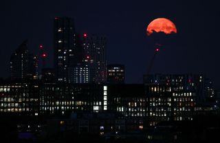 a city scene at night with tall buildings in the foreground and a bright full moon colored orange appearing behind some clouds in the upper right corner of the image.