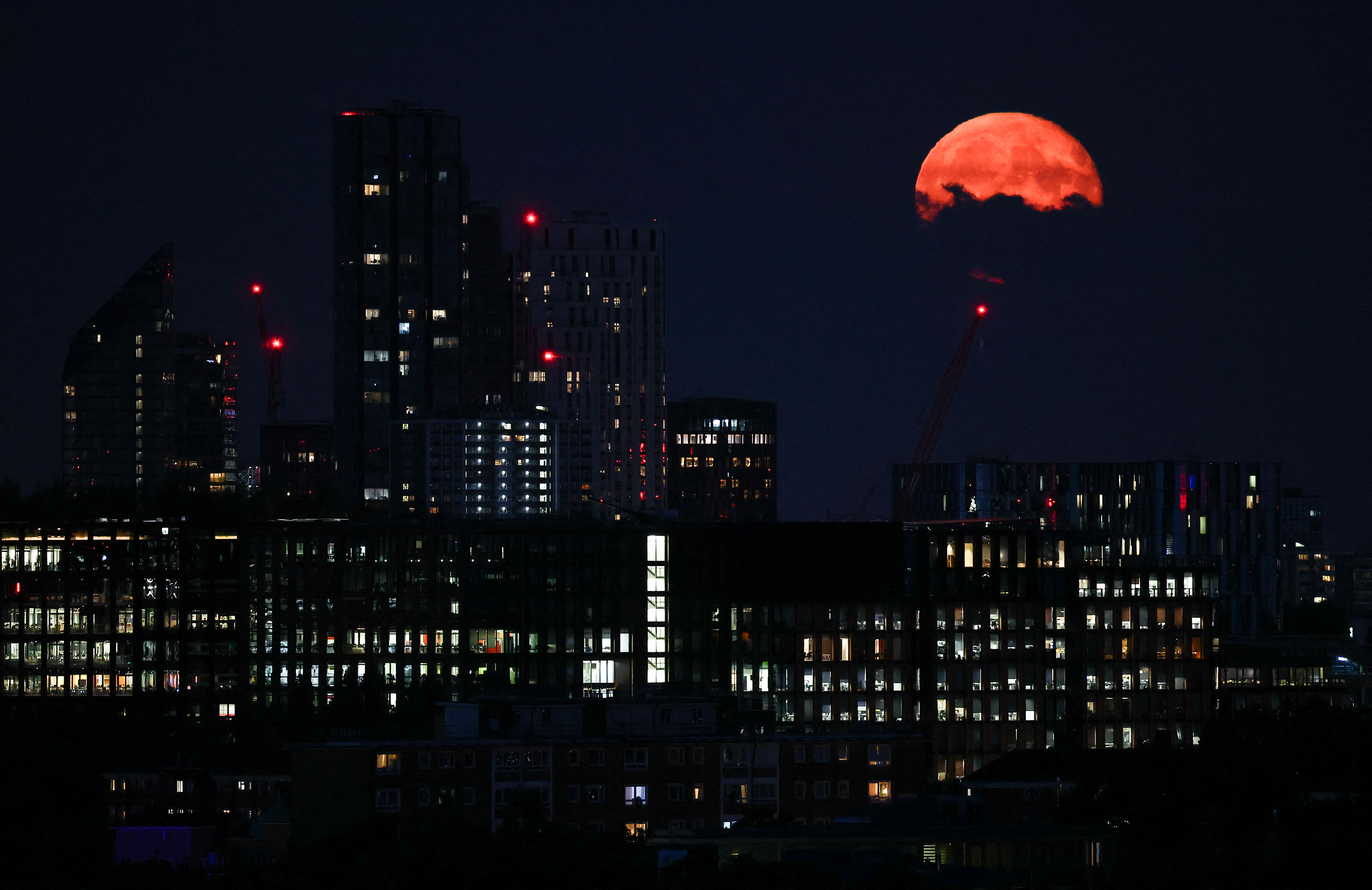 City scene at night with tall buildings in the foreground and a bright orange moon appearing behind some clouds in the upper right corner of the image.