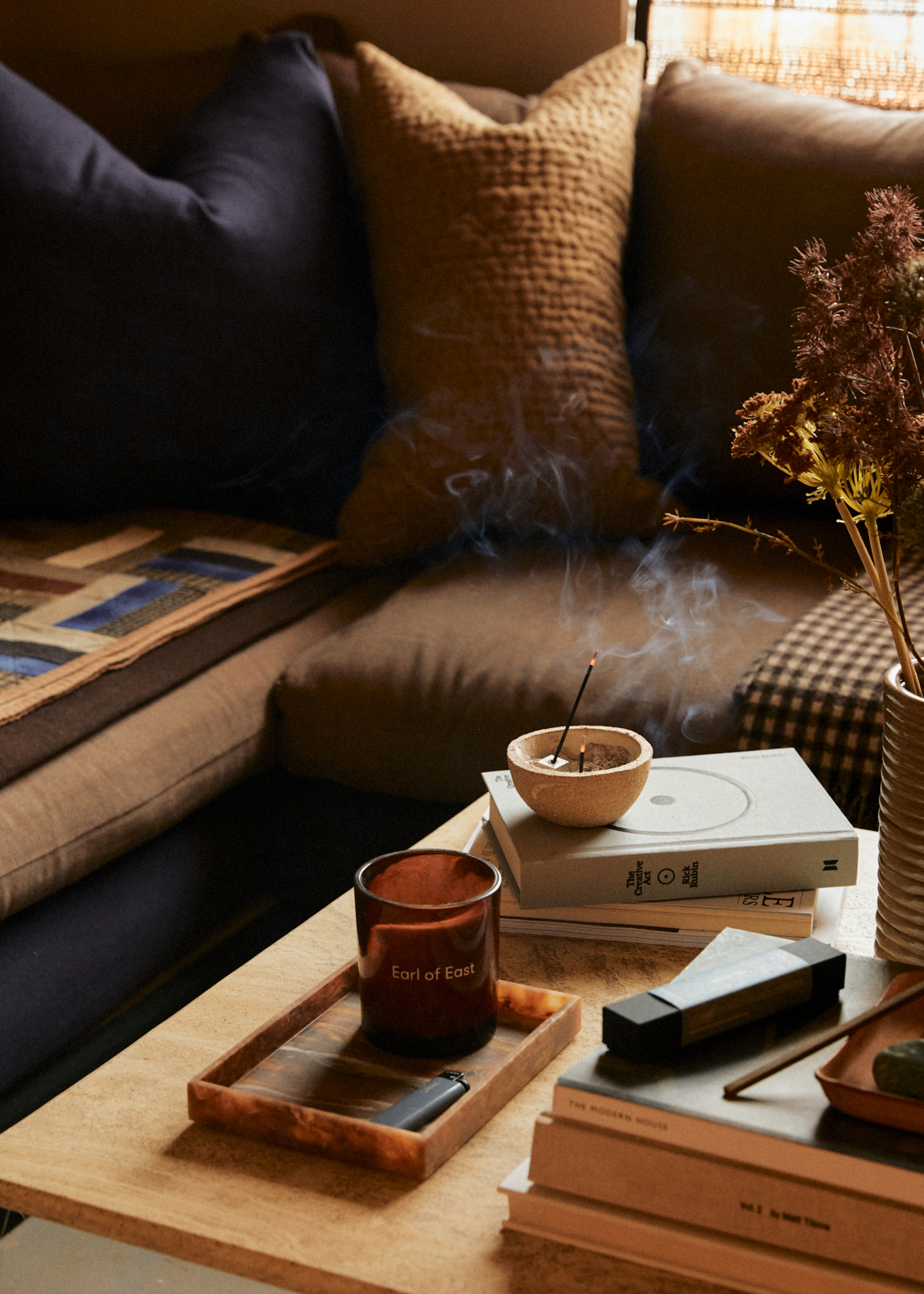 A living room with a wood coffee table featuring a candle in an amber glass and a lit incense stick in an incense bowl on a stack of books