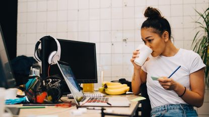 A woman drinking a coffee and working at a desk with a computer