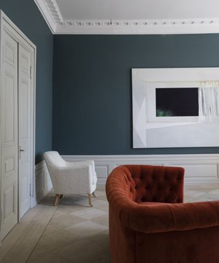 A blue living room with dark blue-green walls, white accents and a burnt orange sofa.