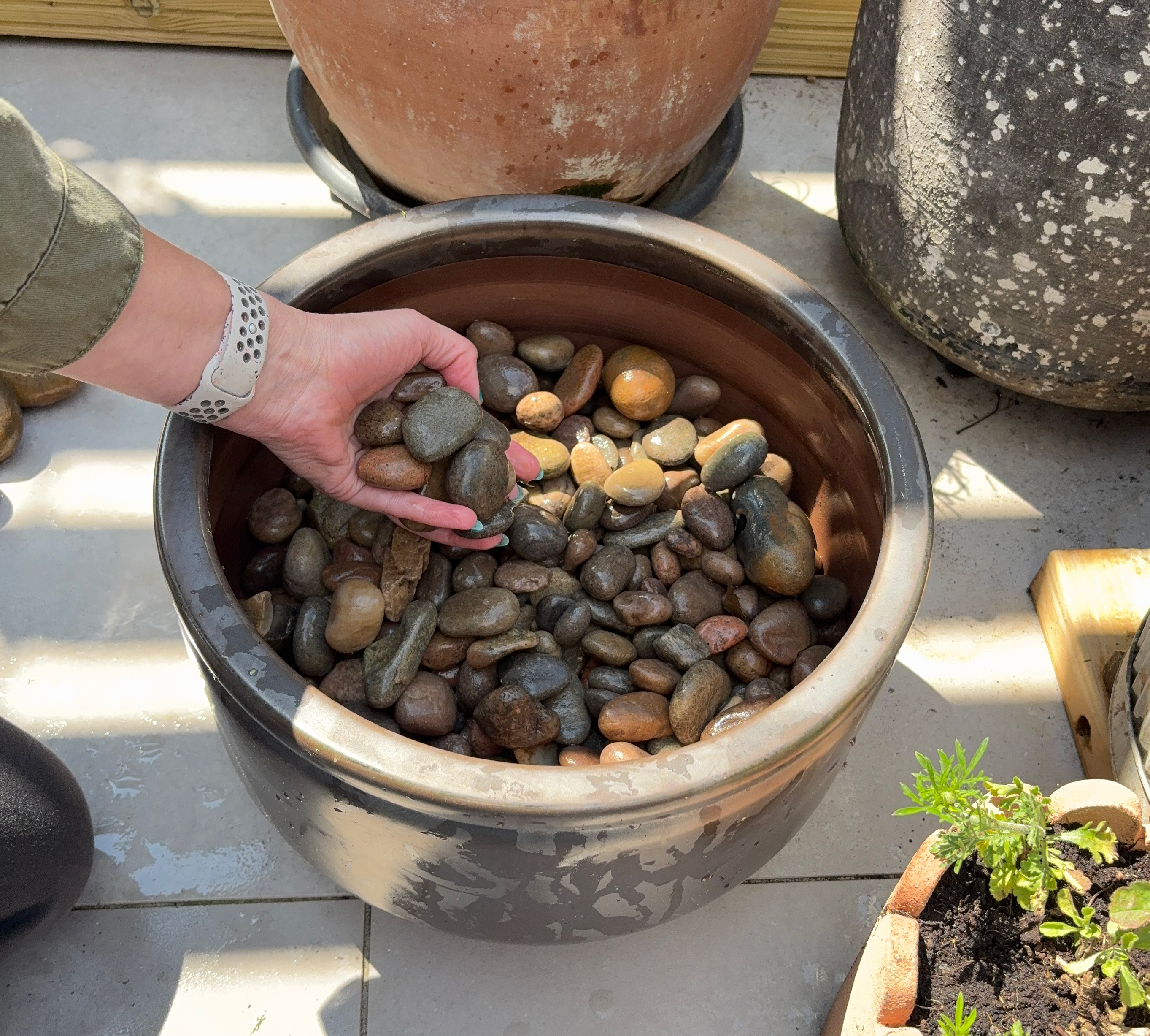 Dark grey circular ceramic planter on a grey patio with a hand filling it with pebbles
