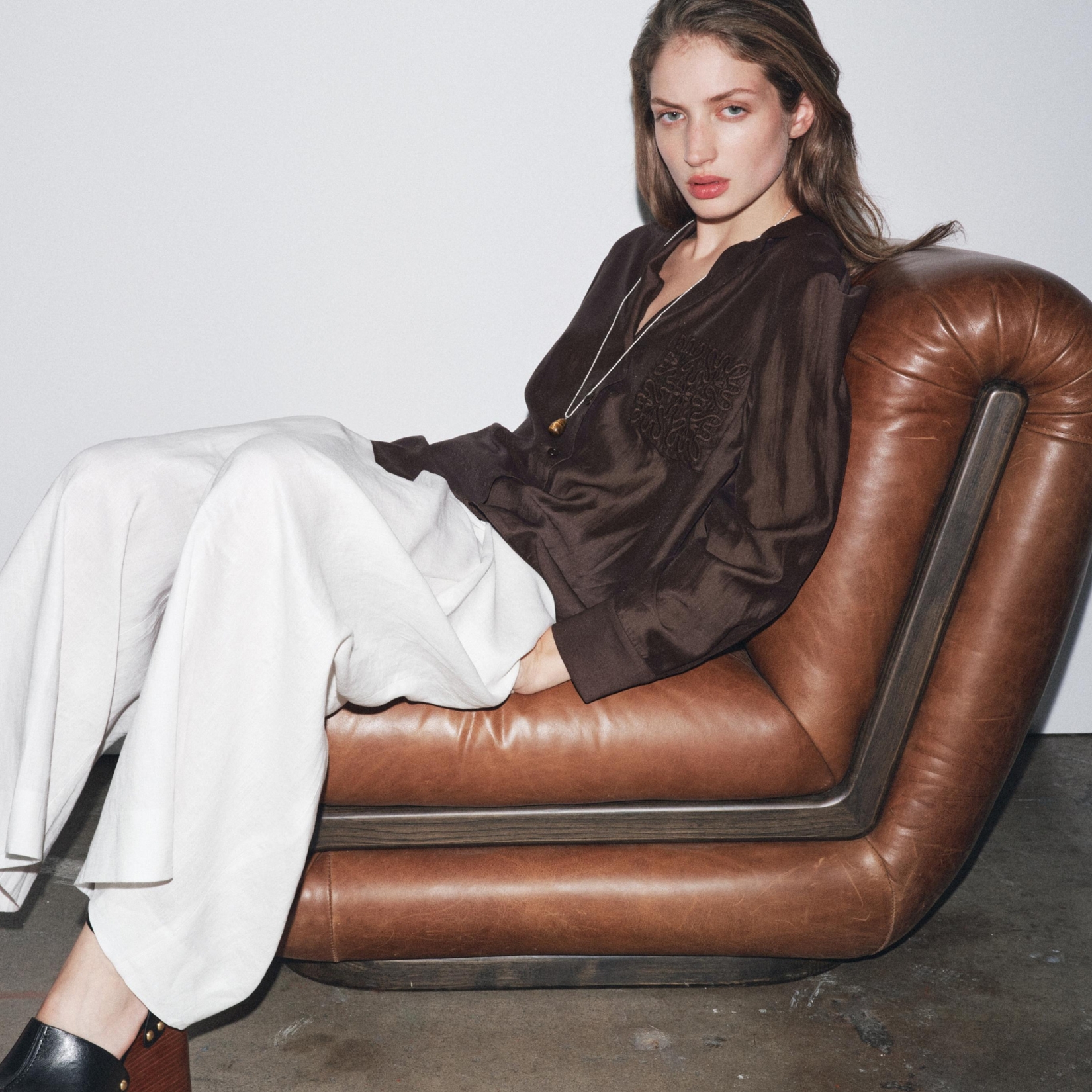 A model wearing a brown blouse and white trousers reclining on a brown leather sofa.