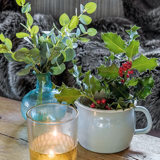 christmas flower arrangements of eucalyptus in a clear blue vase and holly in a blue pot sitting on a wooden table