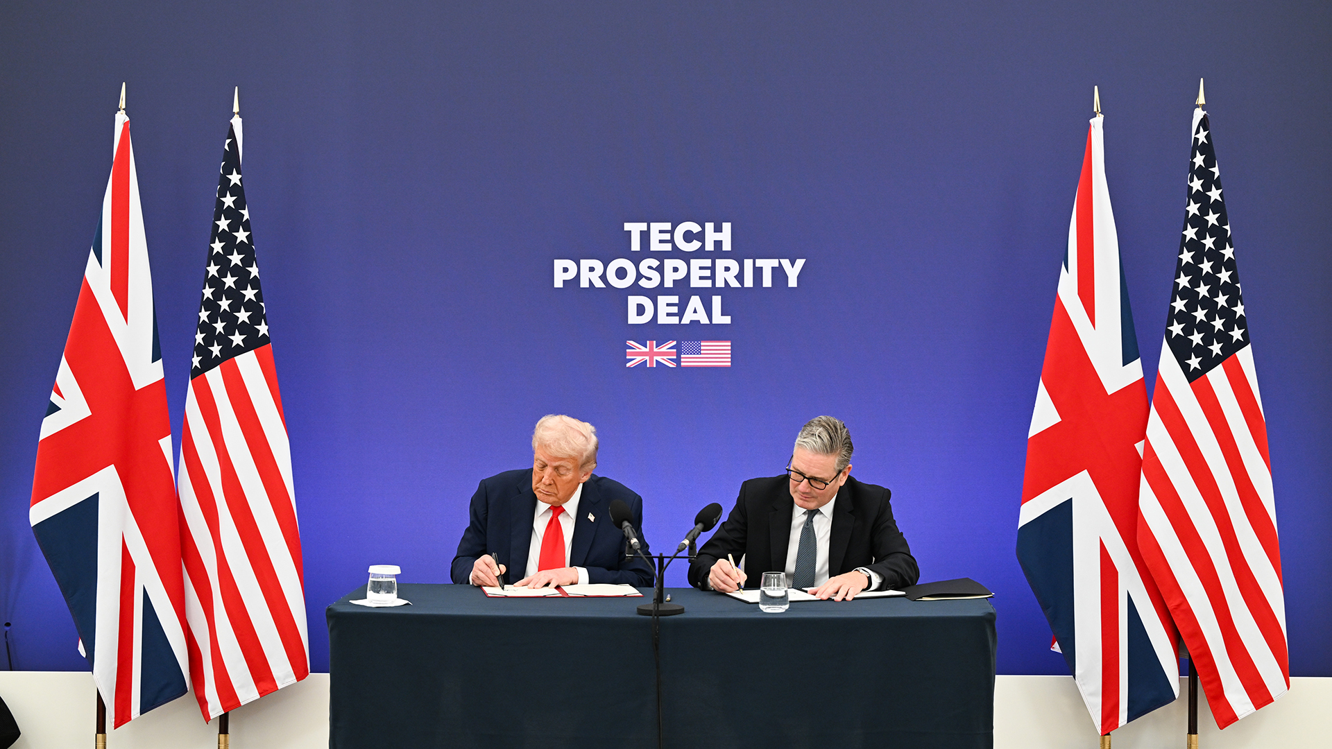 A photograph of US President Donald Trump, on the left, and UK prime minister Keir Starmer, on the right, signing the Tech Prosperity Deal. The pair are sat at a ceremonial table, flanked by UK and US flags. Behind them, a blue wall bears the white words 'TECH PROSPERITY DEAL', with the UK and US flags shown beneath.