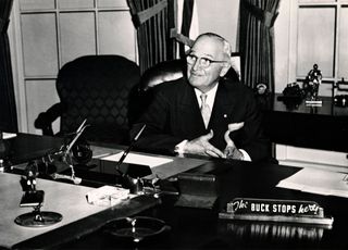 American President Harry S. Truman seated in White House library, with 'The Buck Stops Here' on placard in foreground, circa 1950.