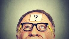 An older man looks up toward a sticky note stuck to his forehead.