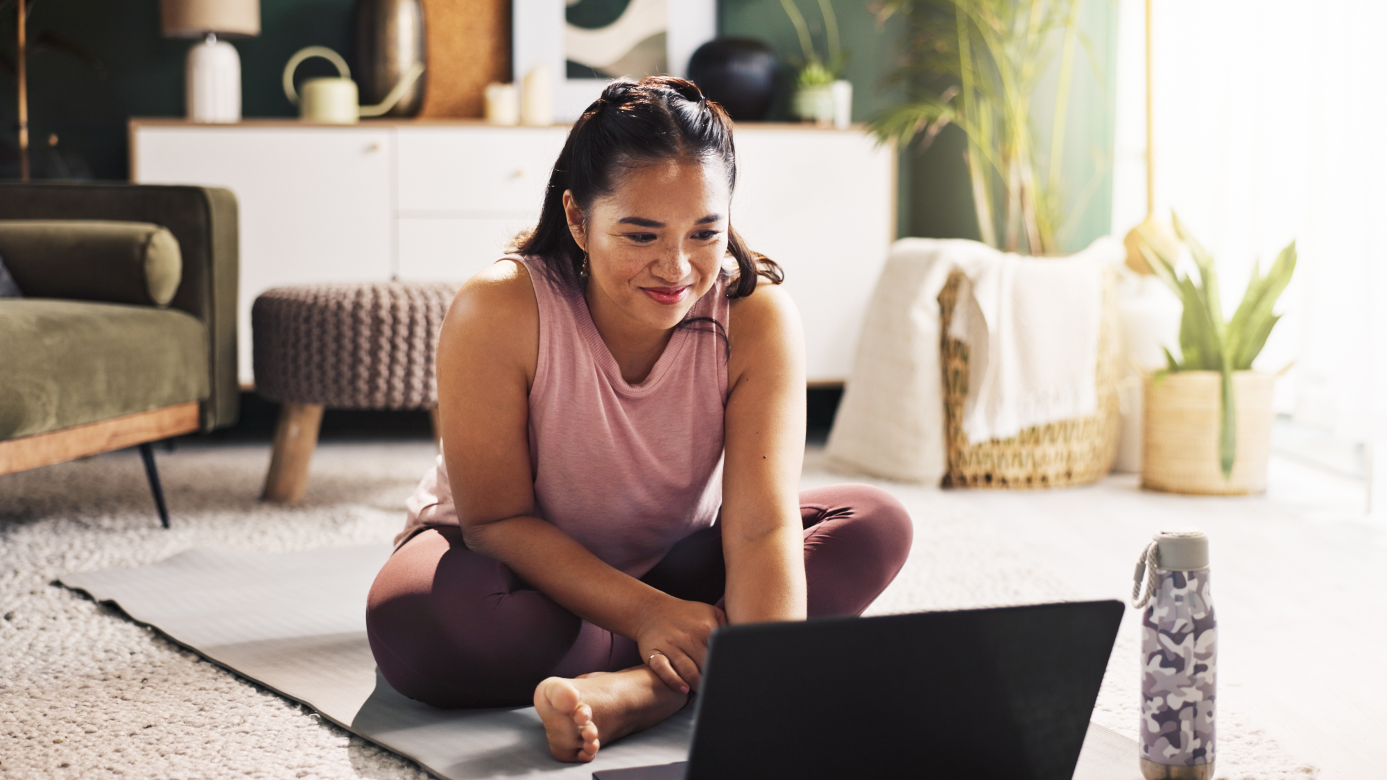 Woman sits cross-legged on yoga mat with open laptop and water bottle on floor in front of her