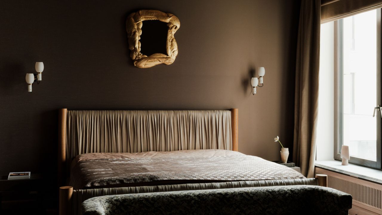 Image of a brown bedroom with a large bed that has a light grey leather headboard. There is a decorative gold mirror hanging above the bed and two unique sconce lights.