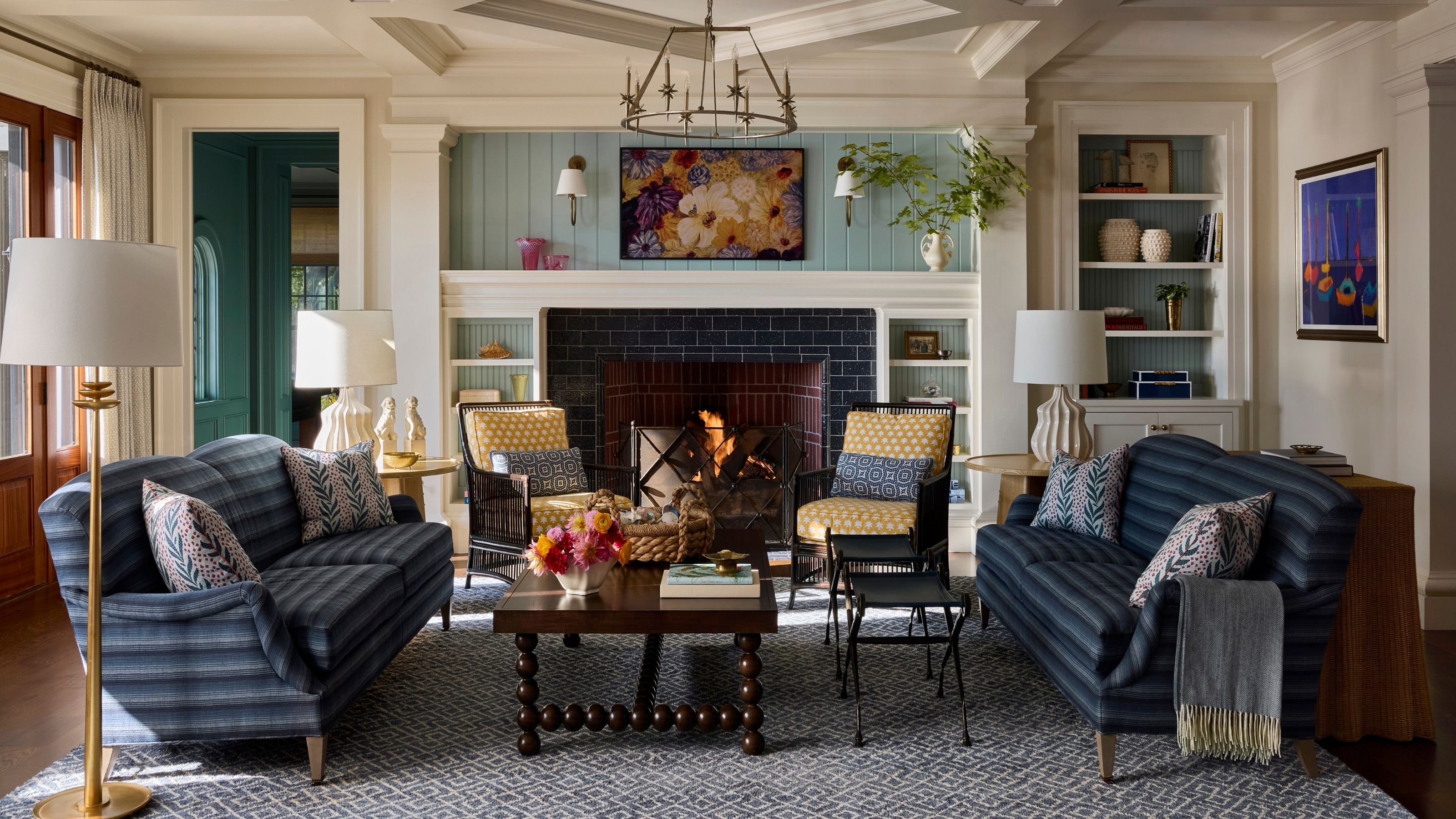 Warm living room featuring twin blue striped sofas, yellow patterned armchairs, a central fireplace with dark tile surround, a low wooden coffee table, and coffered ceilings.