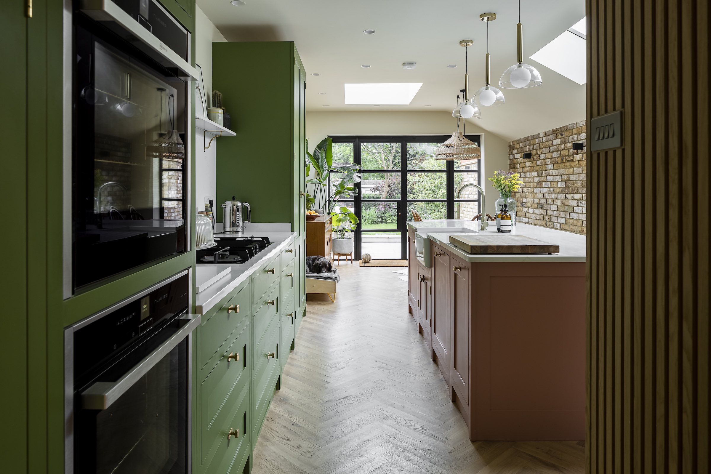 side return extension with pink and green kitchen. The kitchen island is pink and above the island are light pendants hanging. Opposite the island are green kitchen cabinets.