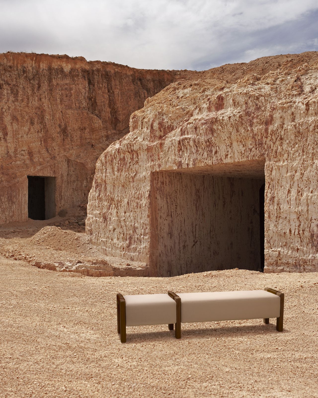 A wood and textile beige-brown banquette sits in front of two reddish rock caves in an empty landscape.