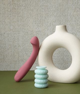 Grey textured background, olive green base, cream ceramic donut shape vase next to a pink and a light blue sex toy