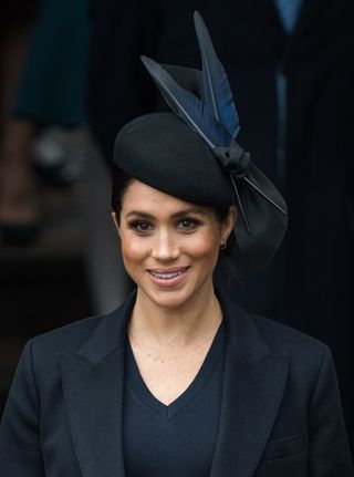 Meghan Markle wearing a black coat and black hat with a big feather smiling on Christmas Day 2018