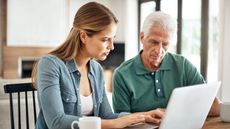 A young woman helps her aging father with his finances on a laptop at the kitchen table.