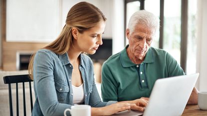 A young woman helps her aging father with his finances on a laptop at the kitchen table.