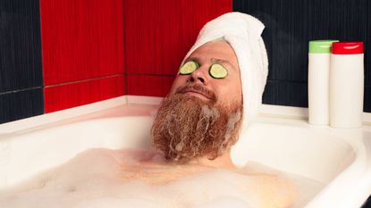 A man with his hair wrapped in a towel and cucumber slices on his eyes relaxes in a bathtub.