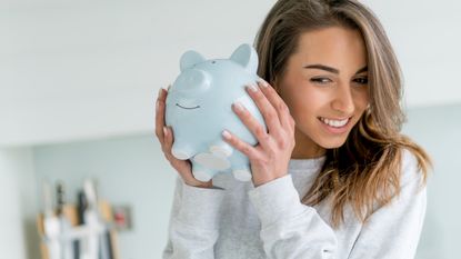 A young woman holds a piggybank up to her ear as if listening to it.