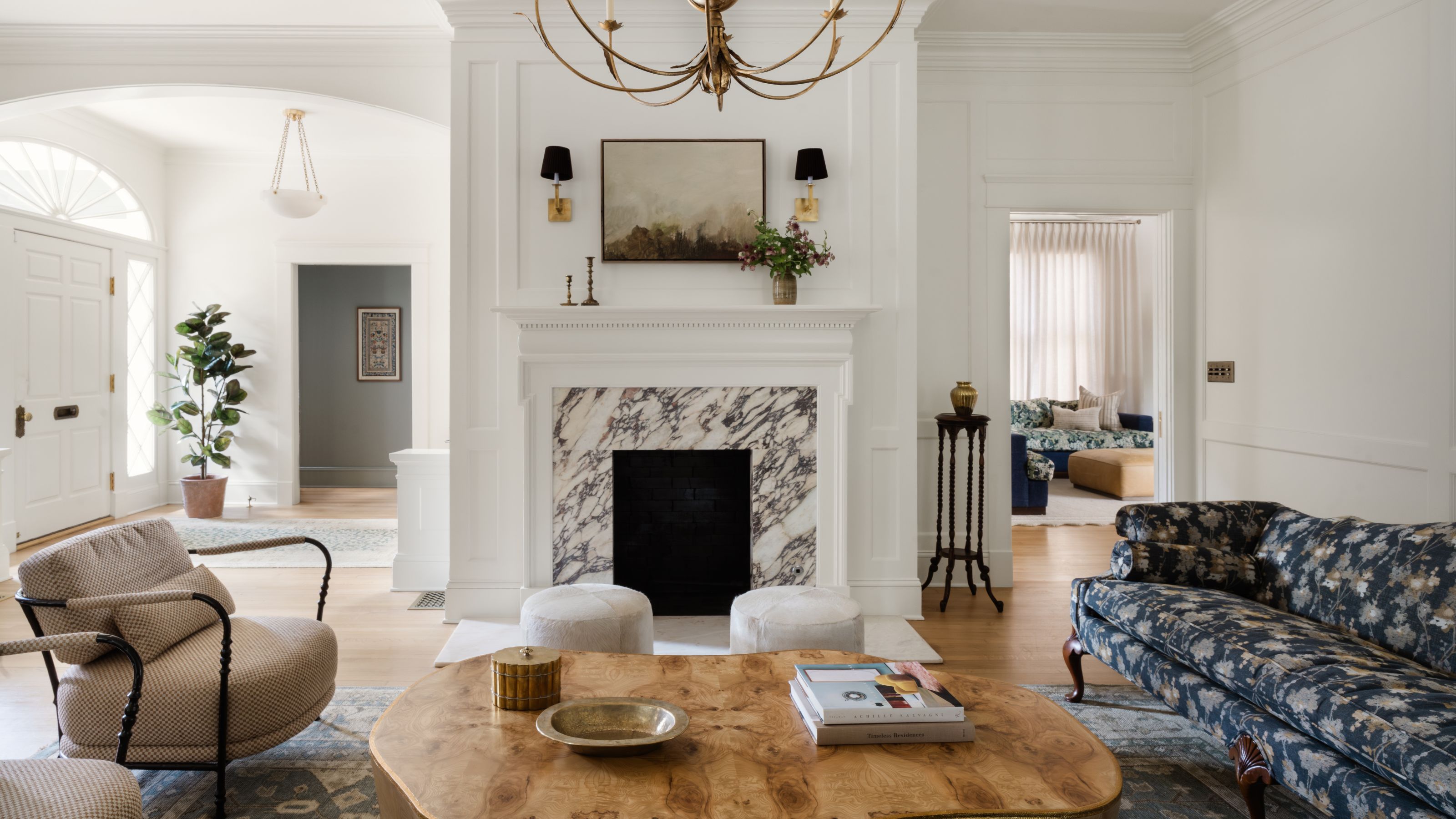 a formal living room with views through to the entryway with a blue floral vintage sofa, marble fireplace, wooden coffee table and collection of modern armchairs and footstools