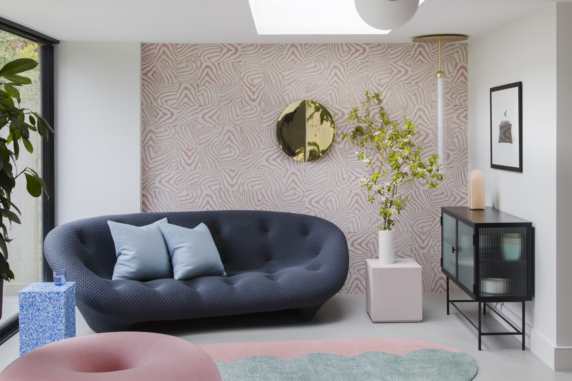 sitting area with pink squiggle wallpaper, skylight, fluted glass cabinet with artwork above, pink side table with vase of flowers, navy round sofa, colorful rug, pink donut floor cushion, and blue speckled side table
