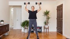 A woman exercises at home. She's holding dumbbells overhead while standing; behind her are green plants and shoes. 