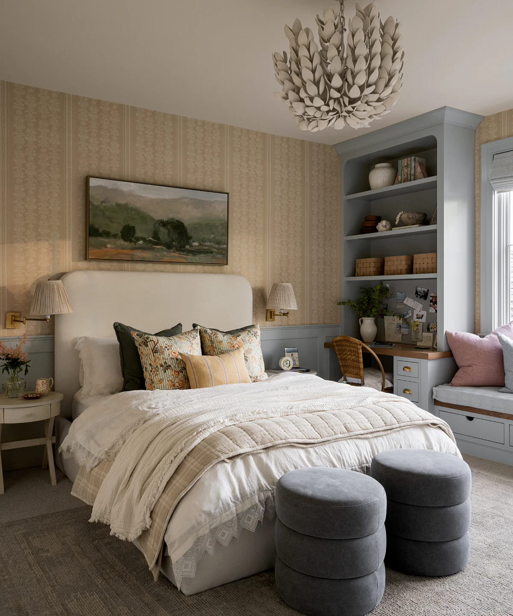 a bedroom with a large bed with white bedding on top, two blue ottomans at the foot of the bed, floral pillows on the bed, a wall of built-in shelving, two sconces on either side of the bed, a chandelier, and a landscape painting above the bed
