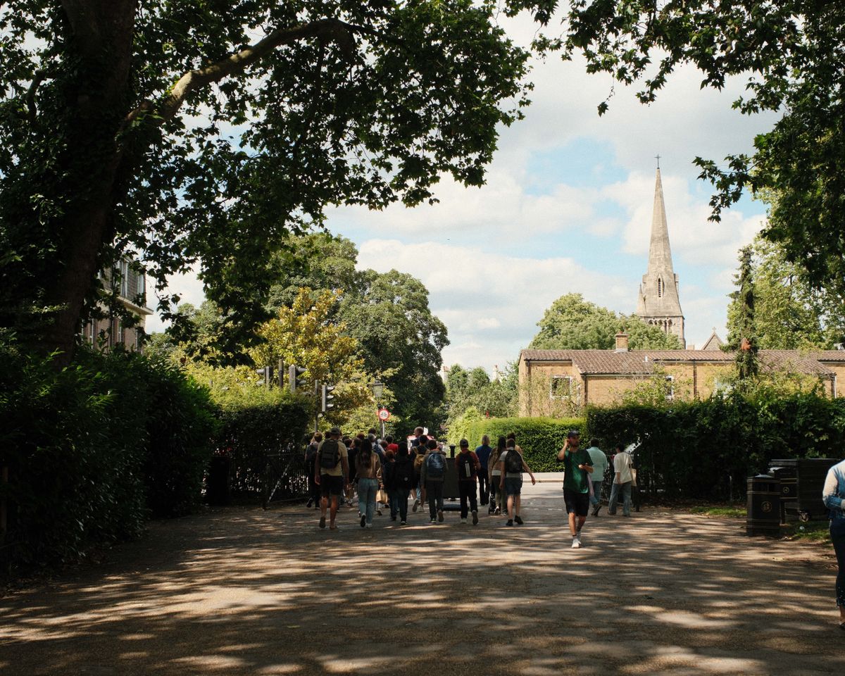 Trees leading to a bridge with lots of people crossing and a church spire in the background