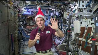 Thomas Pesquet wearing a Santa hat and holding a microphone while waving at the camera.
