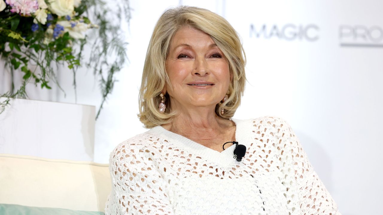 Martha Stewart with a blonde bob wearing a white shirt, with a vase of white flowers and a white backdrop behind it