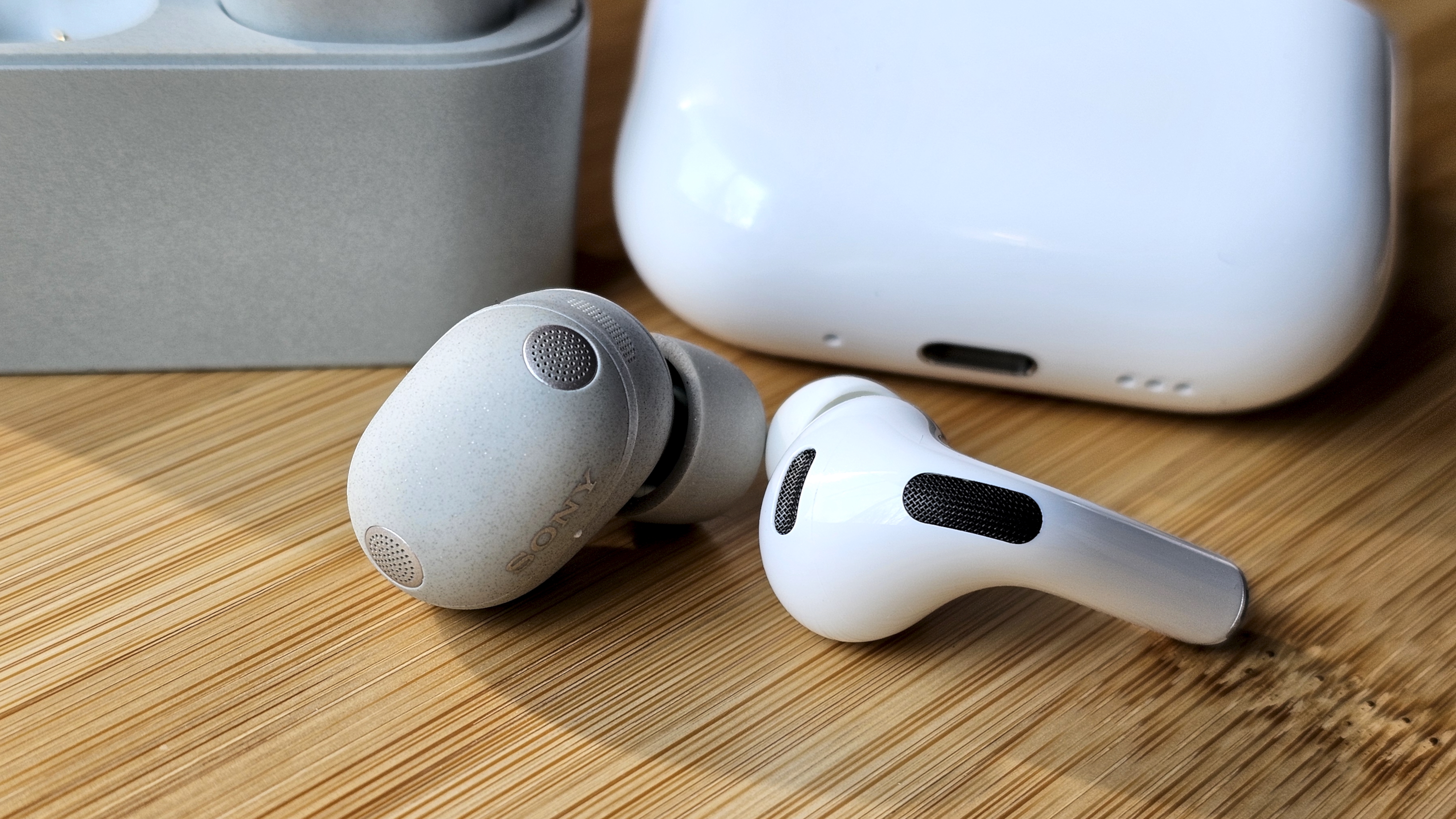 Sony WF-1000XM6 earbuds next to the Apple AirPods Pro 3, on a wooden table, in a versus-style format