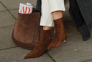 Woman sitting on bench wearing brown suede boots with gray coat and white jeans.