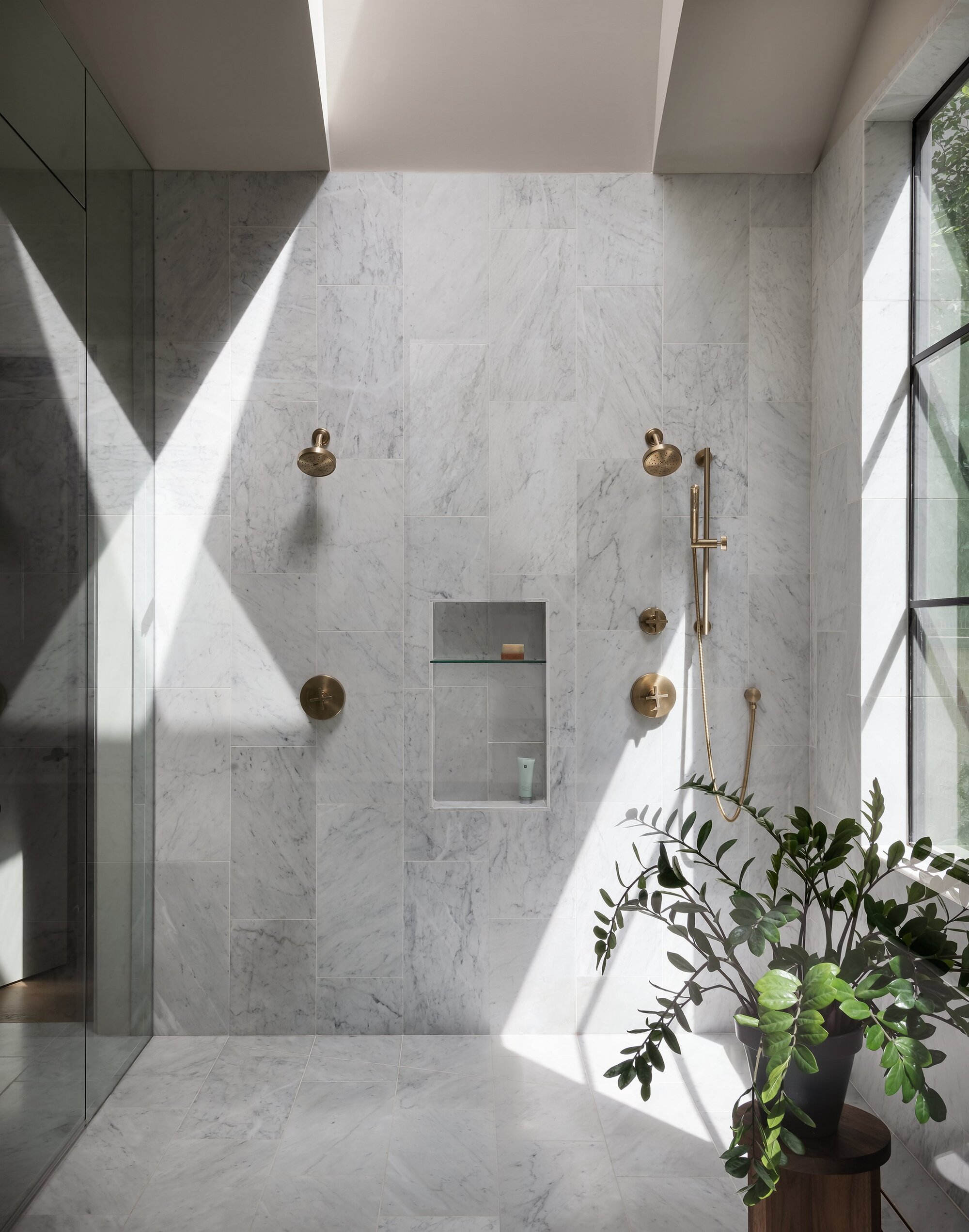 modern bathroom with tiled walls and floors, with a plant in the corner, brass tapware and showerheads, large windows and a skylight letting lots of natural light into the space