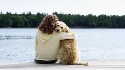 A woman sits on a doc with her arm around her dog.