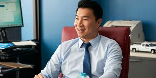 Simu Liu smiles while sitting at his desk in Kim's Convenience
