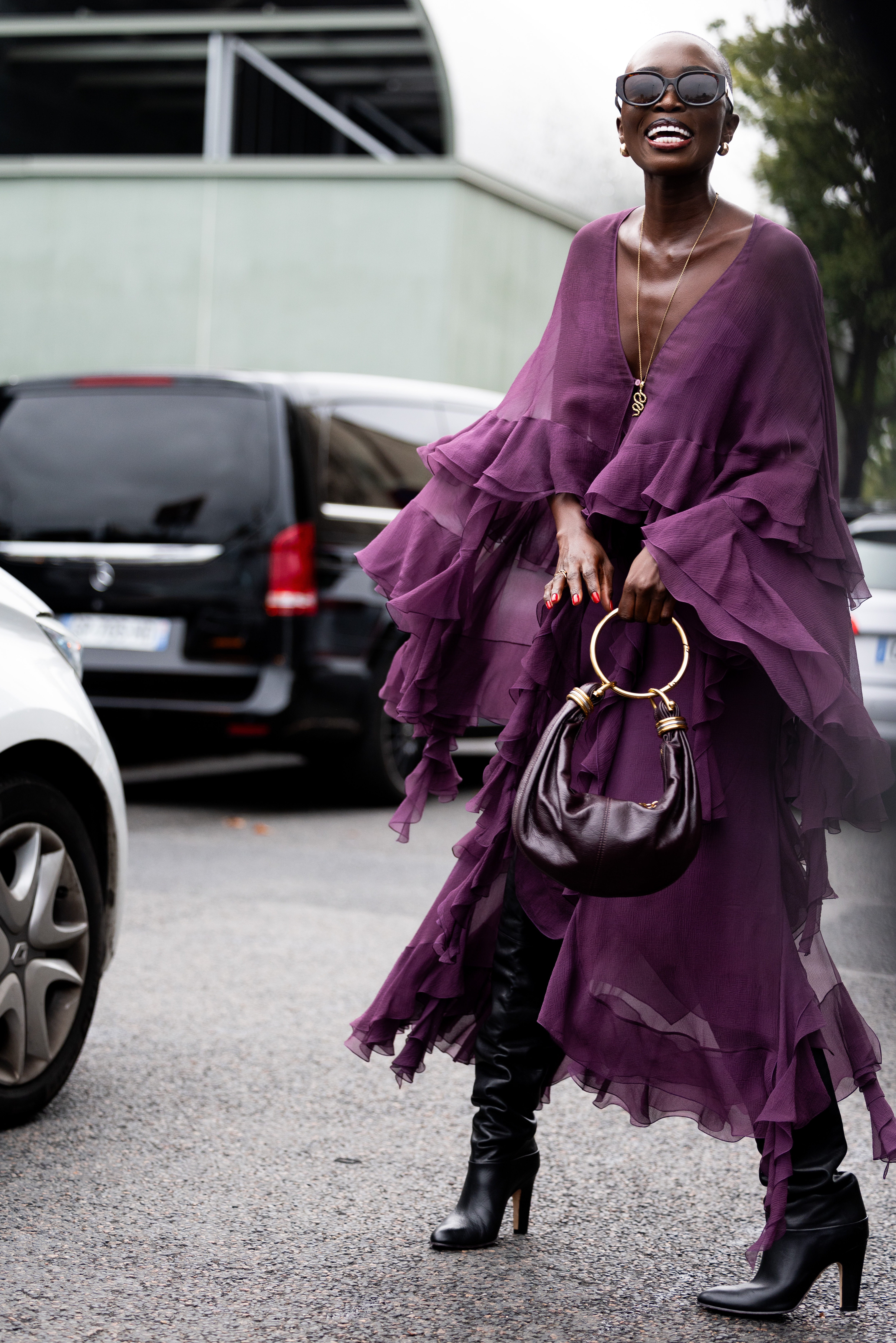 Amina Ladymya lleva un bolso de cuero burdeos y dorado, gafas de sol burdeos, pendientes dorados, un collar dorado, un vestido largo morado de manga larga con volantes y botas de tacón de cuero morado oscuro hasta la rodilla en el exterior del desfile de Chloé durante la Semana de la Moda de Mujer Primavera/Verano 2025.