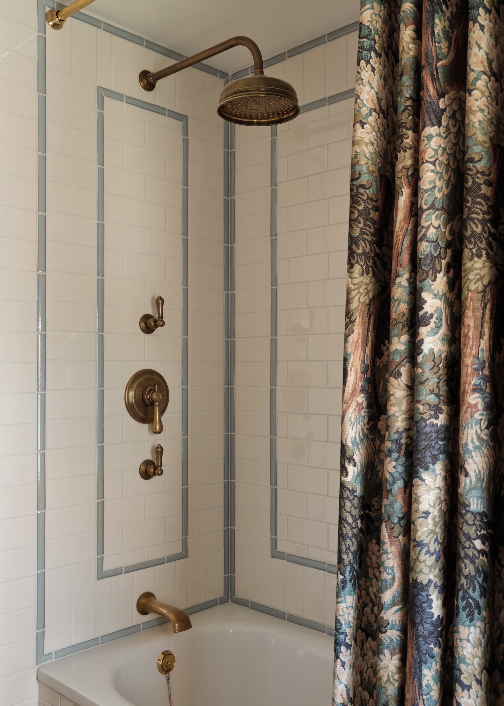 a white tiled bath/shower with antiqued brass hardware and a thick, patterned shower curtain