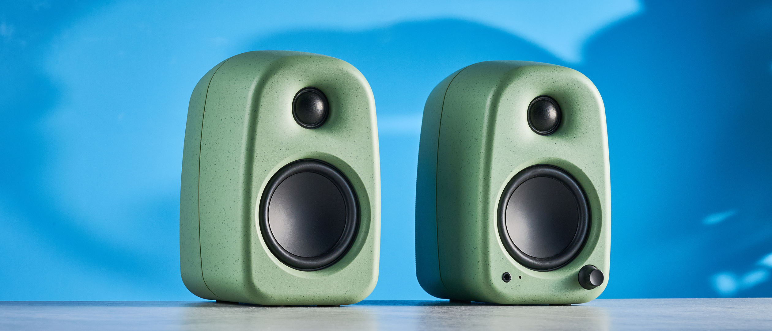Kanto UKI bookshelf speakers in sage photographed in front of a blue background