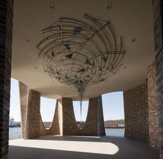Fjordhvirvel, 2018, by Olafur Eliasson installed at the Fjordenhus, Vejle, Denmark. Outdoor area surrounded by a lake with brick archways and a large roof chandelier.