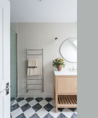 A white door opening up to a bathroom with white tiles on the wall, a silver towel rack with beige towels on, and a wooden vanity unit with a white counter with a vase of flowers and a circular mirror on it