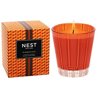 An orange glass jar with an orange candle inside, lit. Anorange patterned cardboard box beside it with a black label on it