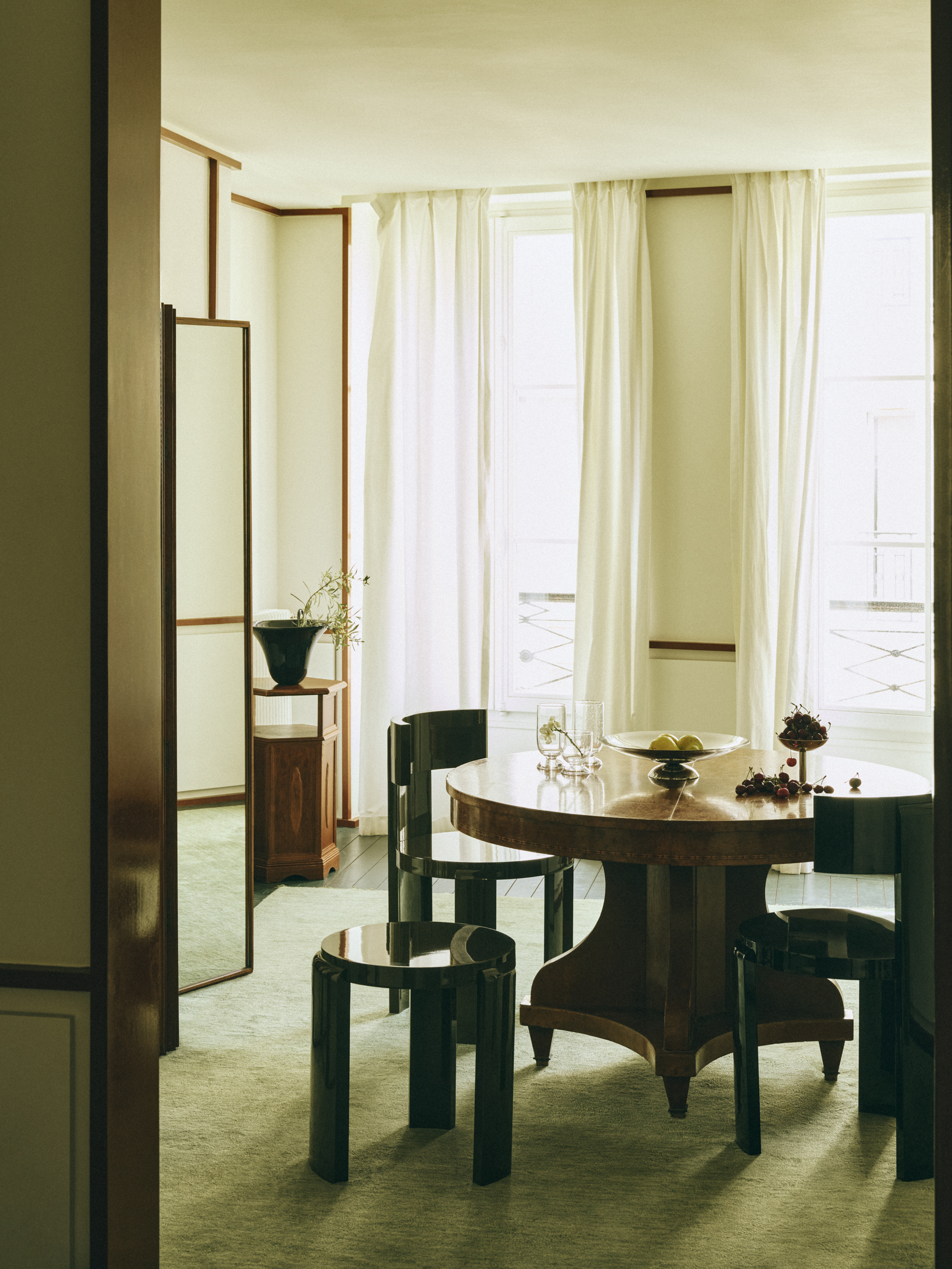 bright room with green carpet, round timber dining table, black chairs, black stool, white curtains, side table with vase