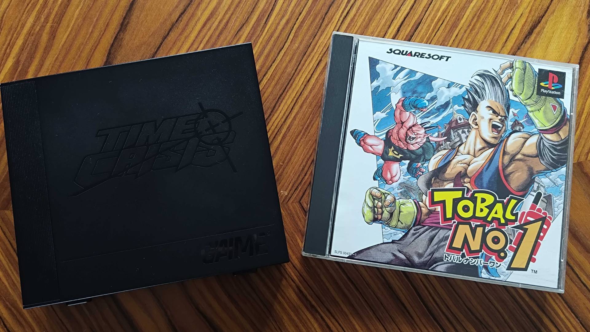 G'AIM'E Time Crisis console next to PS1 Tobal No 1 box on woodgrain table.