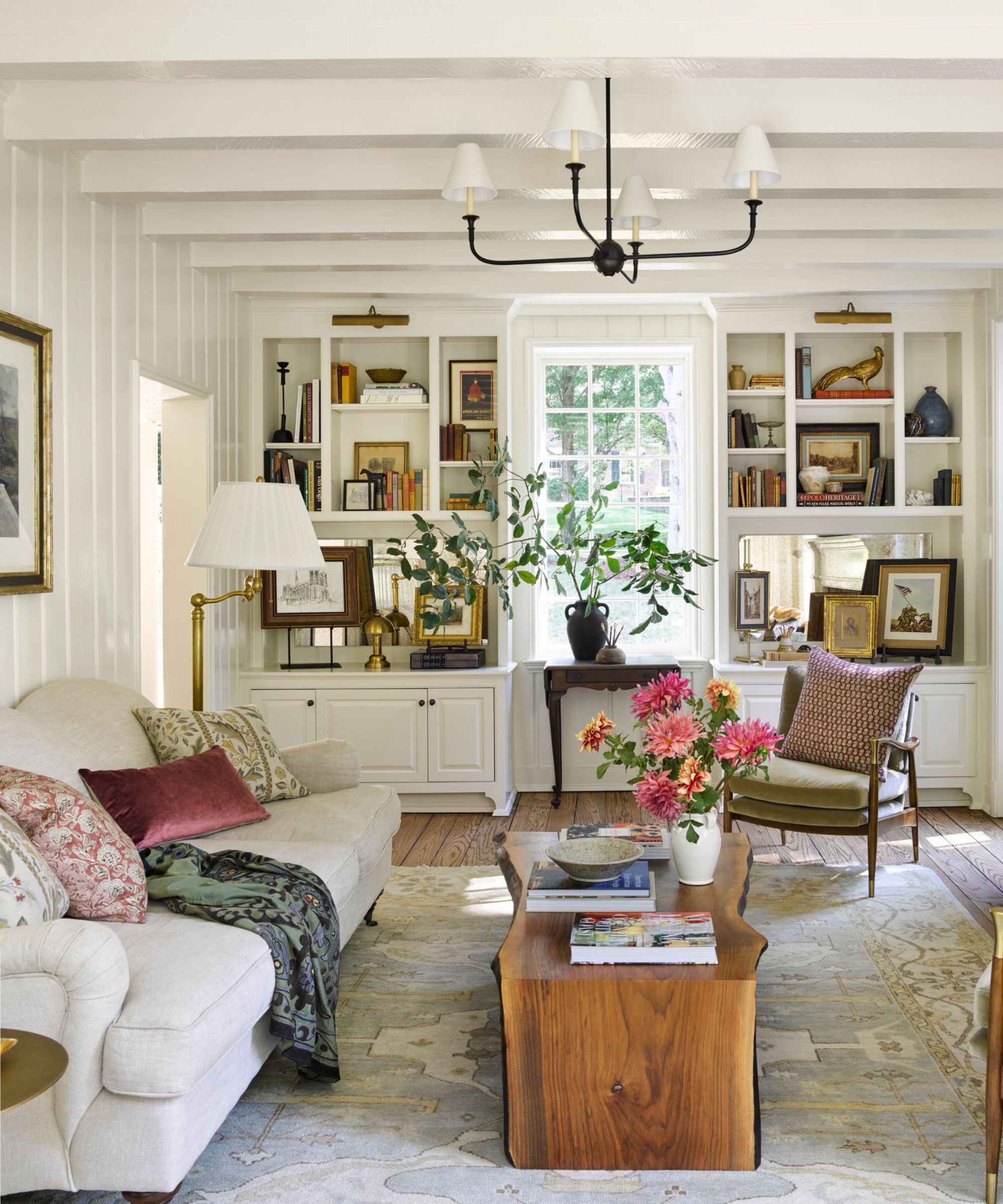 rustic white living room with painted ceiling beams and shiplapped walls, bookcases around the window filled with vintage treasures, a white couch with patterned pillows and throws and a wooden coffee table