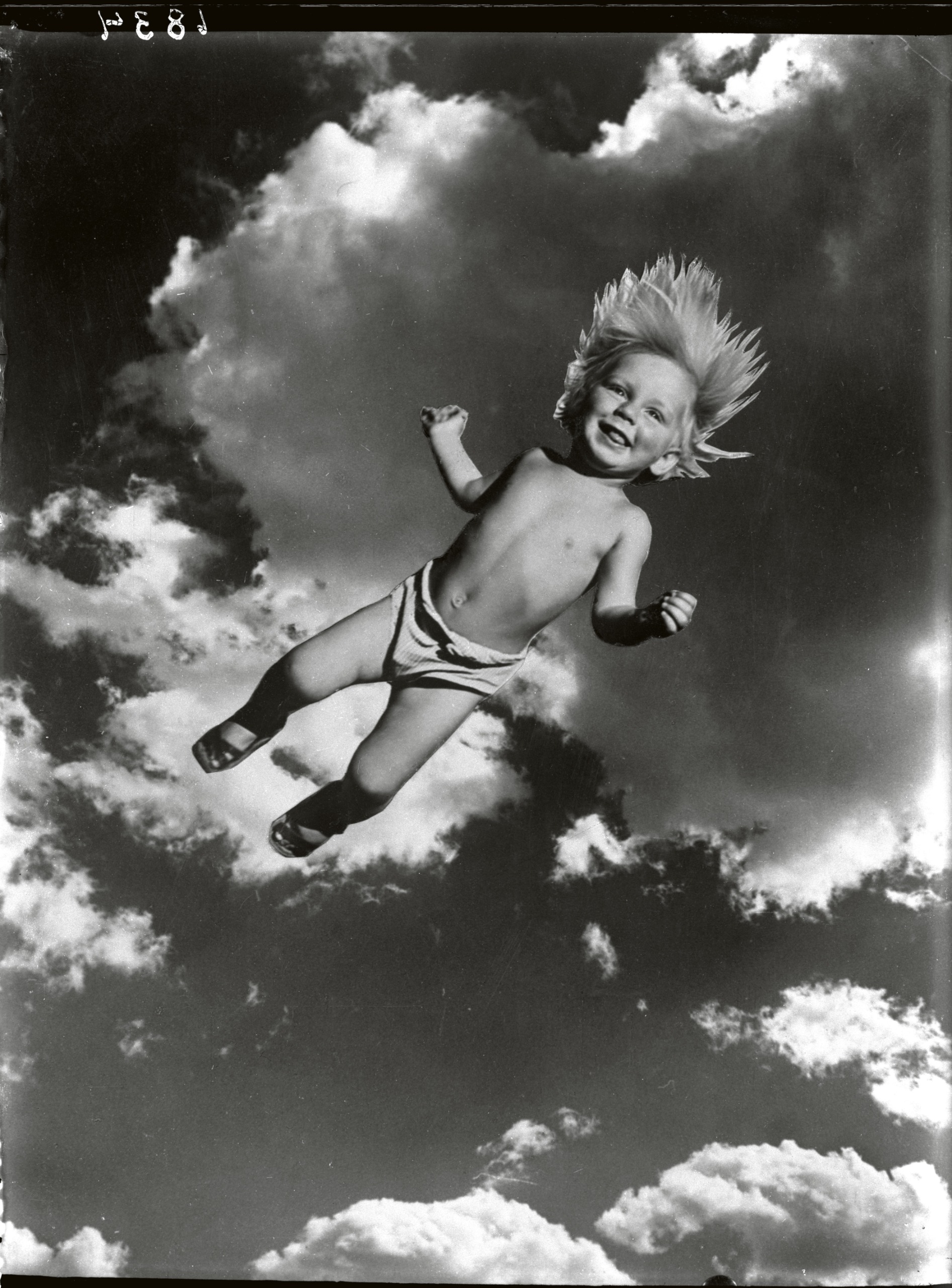 A grinning toddler with wind-blown hair appears to fly through a cloudy sky, photographed from below against a dramatic sky.