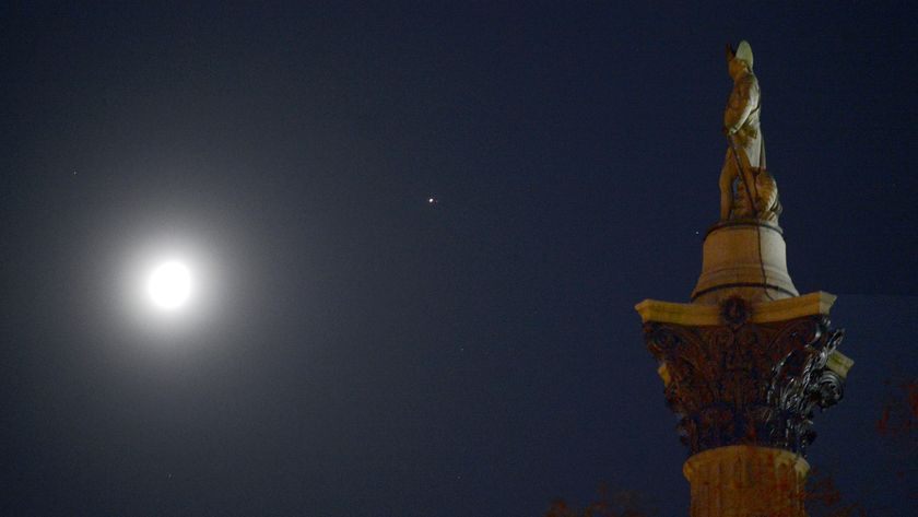 The moon is pictured shining in the night sky with a bright point of light representing Jupiter to its upper left. A stone statue of a military figure atop an ornate column is positioned to the right of the shot, appearing to face the solar system pair.