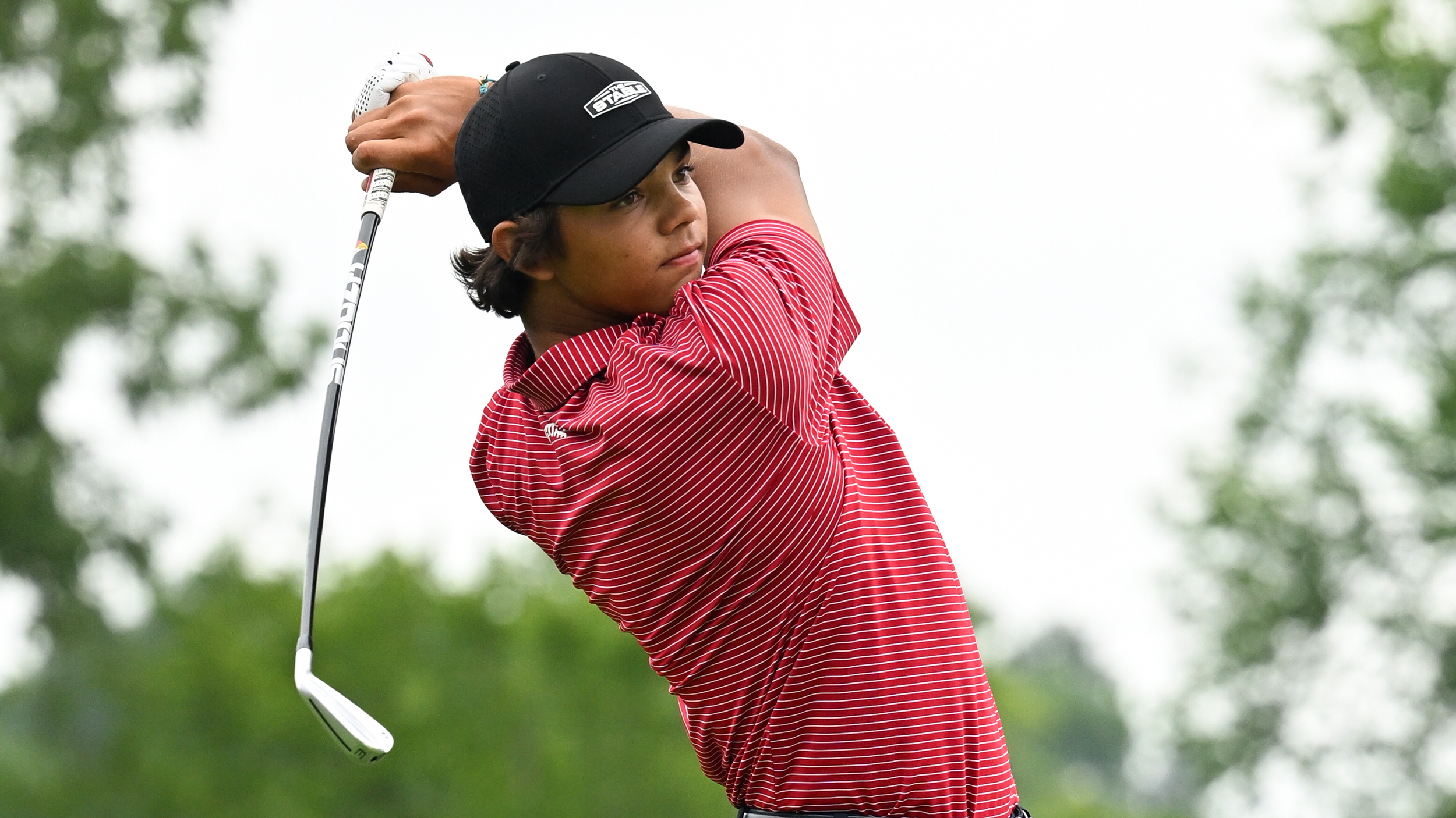 Charlie Woods wearing a red polo and black cap hits an iron shot