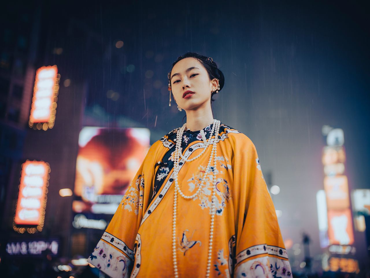 A woman stands confidently in a vibrant orange kimono adorned with intricate patterns, surrounded by colorful city lights in the rain